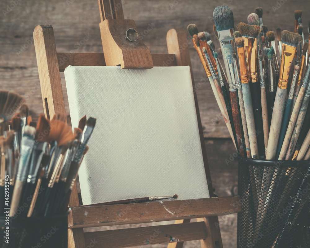 Artistic equipment: empty artist canvas on easel and paint brushes in a artist studio. Retro toned photo.