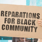 The phrase " Reparations for black community " on a banner in men's hand with blurred background. Demand. Dishonest. Payment. Money. Slaves. Black people. Social inequality. Protest. Tough life